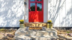 A red door with a welcome mat, white siding, and two potted plants on the steps.