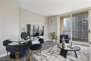 A modern living room with a glass table, dark upholstered chairs, a round coffee table, and a large TV on the wall.