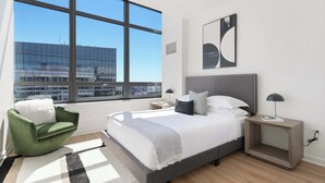 A modern bedroom with a large window, a green chair, a bed with white bedding, and a nightstand with a lamp.