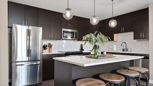 Une cuisine moderne avec des meubles de couleur sombre, des appareils en acier inoxydable, un îlot central doté de tabourets et un éclairage suspendu.