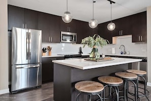 A modern kitchen with dark cabinetry, stainless steel appliances, a central island with stools, and pendant lighting.