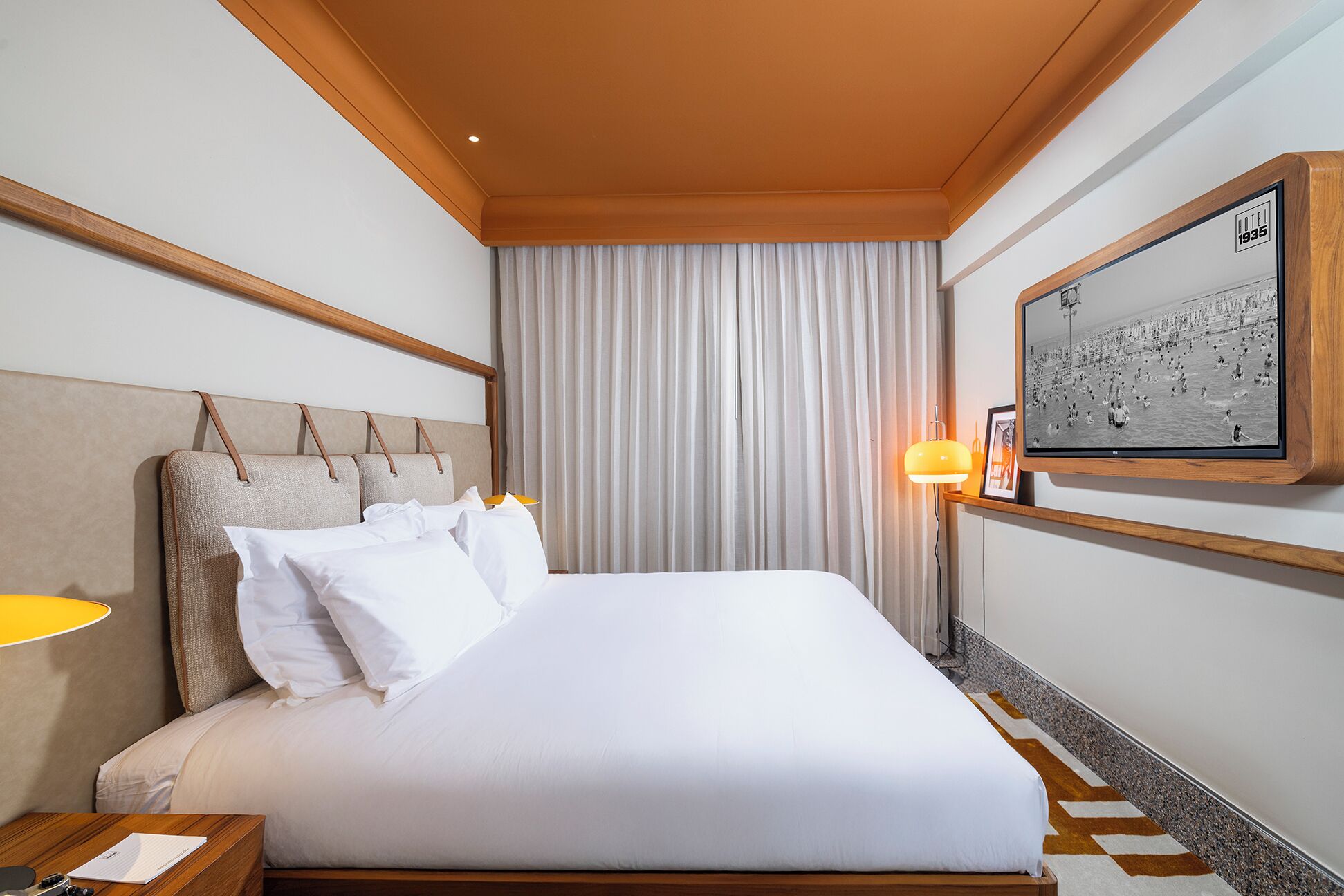 A hotel room with a large bed, a headboard with multiple pillows, a nightstand with a lamp, a framed picture on the wall, and a window with curtains.