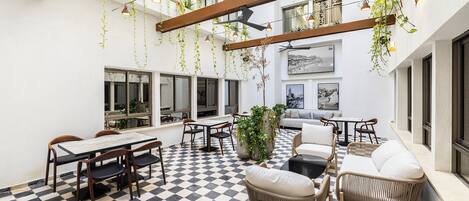 A spacious indoor area with checkered flooring, wooden tables and chairs, hanging plants, and large windows providing views of the city.