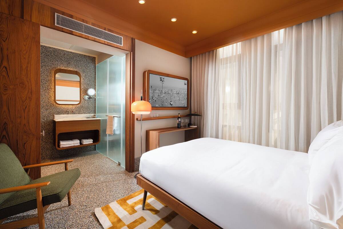 Ein Hotelzimmer mit Bett, Holzstuhl, kleinem Schreibtisch mit Lampe, Spiegel und Bad mit Waschbecken und Handtuchhalter.