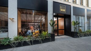 Une entrée d’hôtel moderne avec une grande façade vitrée, un espace extérieur aménagé avec des plantes et une enseigne indiquant « HÔTEL 1935 ».