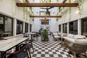 Un comedor interior con suelo a cuadros, mesas y sillas de madera. Hay plantas colgantes y un ventilador de techo.