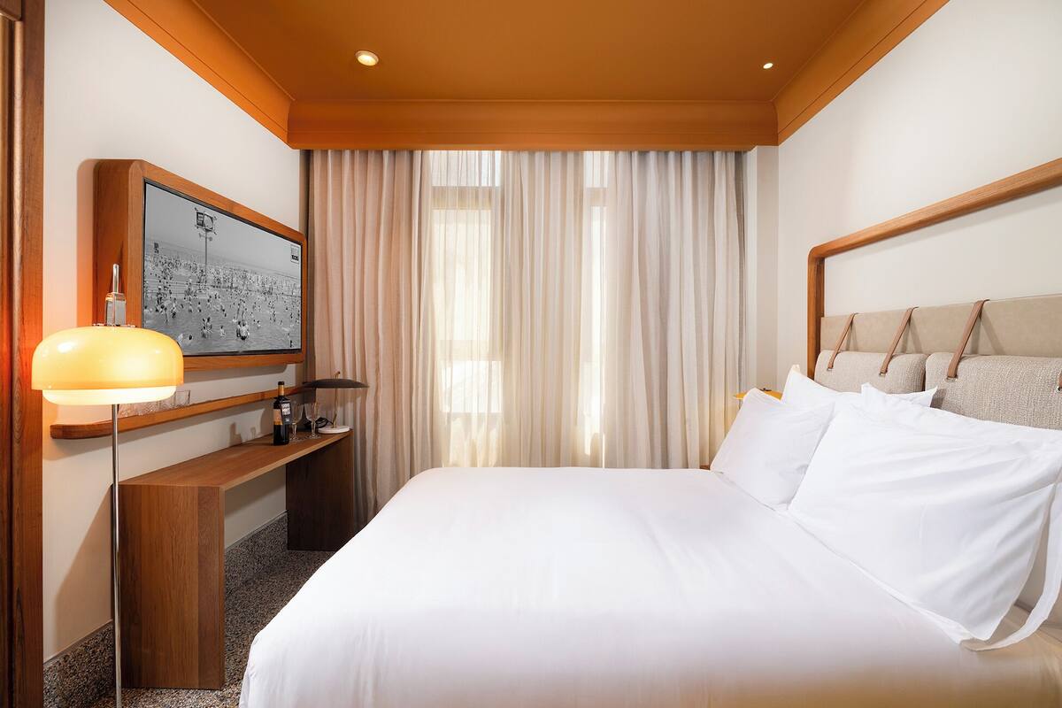 Ein Hotelzimmer mit einem großen Bett, einem Schreibtisch mit Lampe, einem Fernseher und einem Fenster mit Vorhängen.