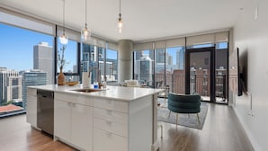 Una cocina moderna con una isla grande, un comedor con una silla color aguamarina y vistas a la ciudad a través de amplios ventanales.