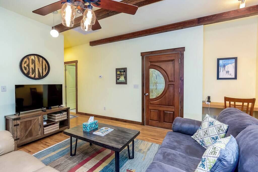 A living room with a sofa, a coffee table, a TV, wooden beams, a ceiling fan, and a door.