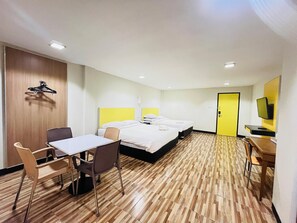 Quarto de hotel com duas camas, uma escrivaninha, cadeiras, televisão e chão de madeira.