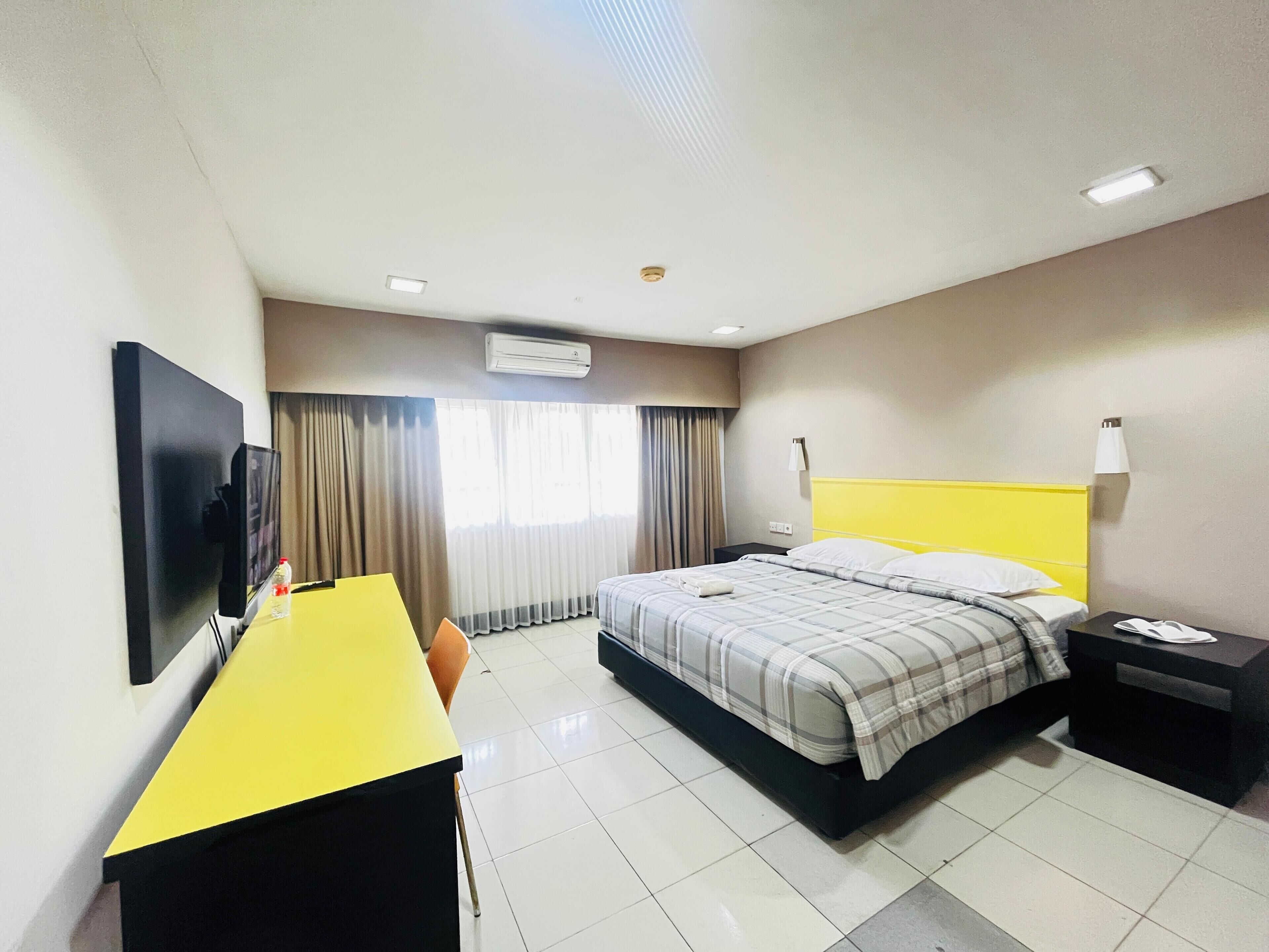 A hotel room with a bed, a desk, a chair, a TV, and an air conditioning unit.