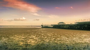 Un muelle se adentra en el agua, con un edificio al final, una playa de arena y un cielo despejado.