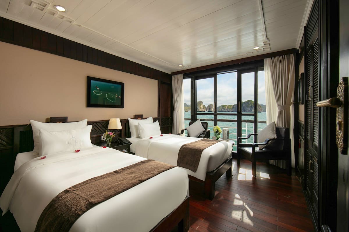 A hotel room with two beds, a large window with a view of the sea, a table with chairs, and a painting on the wall.