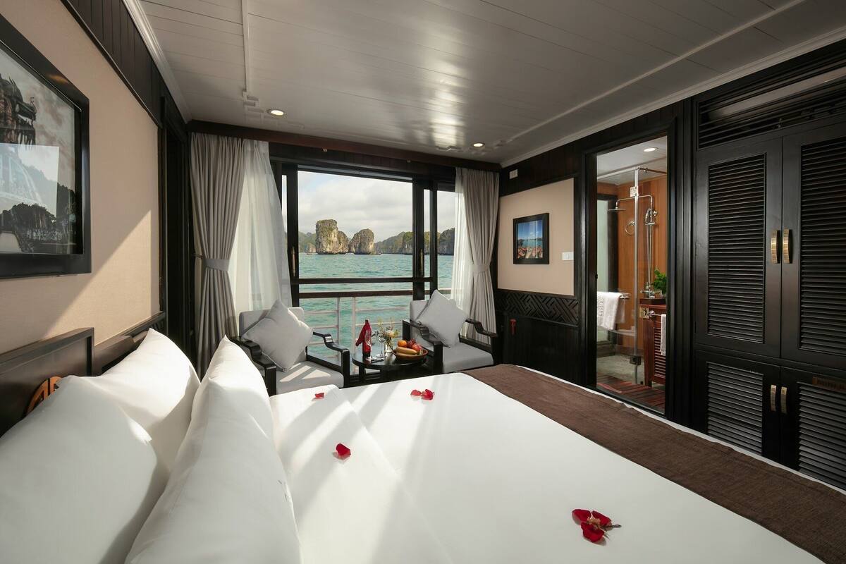 A hotel room with a large bed, two armchairs, a small table with fruit and flowers, and a view of the sea.