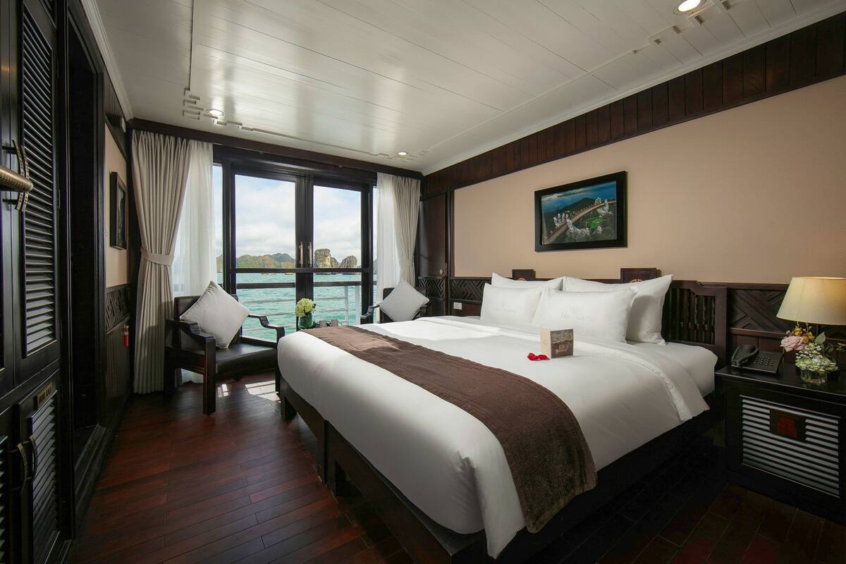 A hotel room with a large bed, a desk, a chair, a nightstand, and a window with a view of the sea.