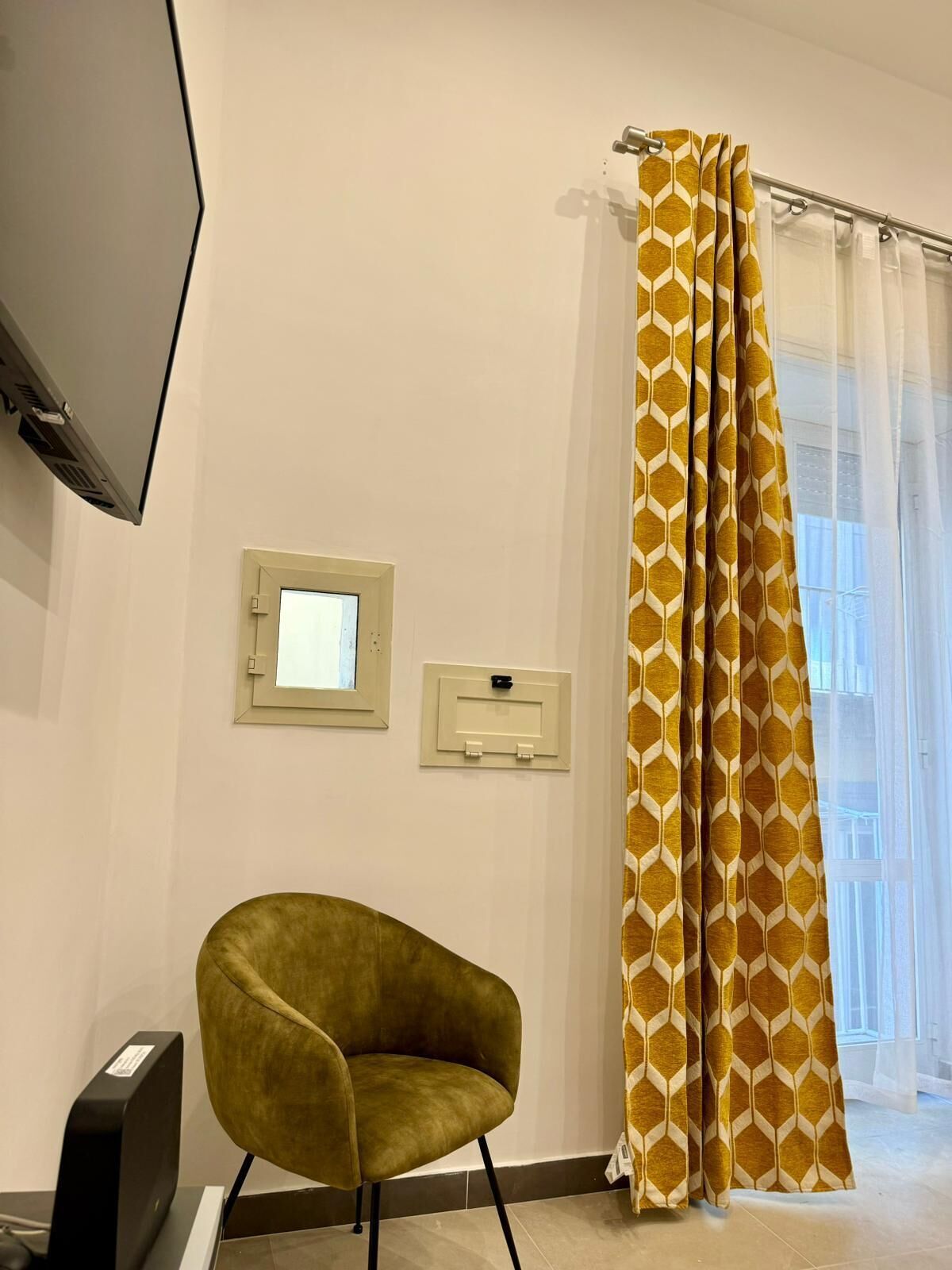 A room with a single green armchair, a flat-screen TV mounted on the wall, a window with a curtain, and a wall-mounted light switch.