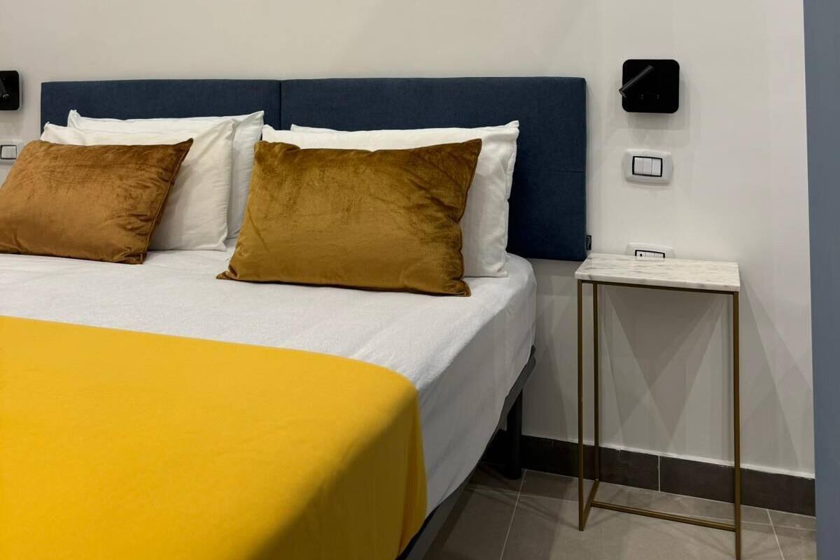 Una cama bien hecha con una colcha amarilla, dos almohadas marrones y un cabecero azul. Hay una mesita de noche pequeña con una lámpara y un interruptor de luz en la pared.