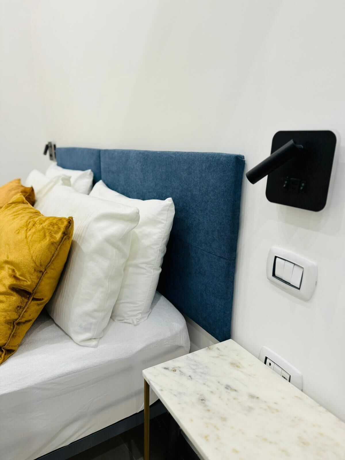 A bed with white and yellow pillows, a blue headboard, a small table with a marble top, and a wall-mounted switch and socket.