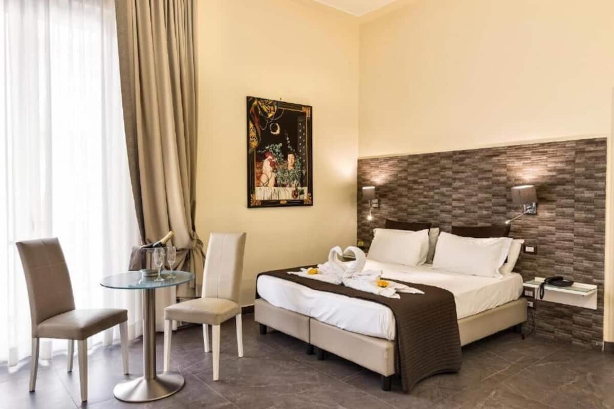 Habitación de hotel con una cama, dos sillas, una mesita redonda pequeña, una mesita de noche y una lámpara de pared.