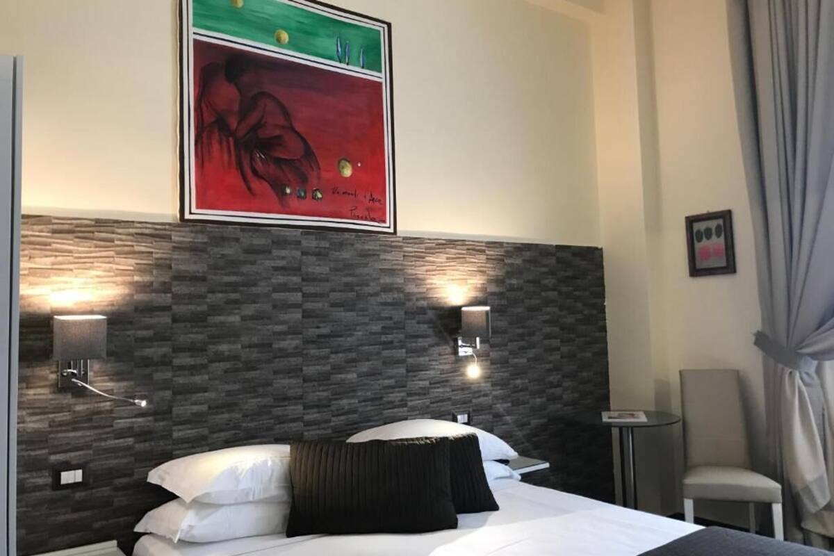 Habitación de hotel con diseño moderno, que incluye una cama grande con ropa blanca, un cabecero texturizado oscuro y dos lámparas de lectura fijadas a la pared.