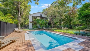 A modern house with a swimming pool, surrounded by lush greenery and a wooden deck with lounge chairs.