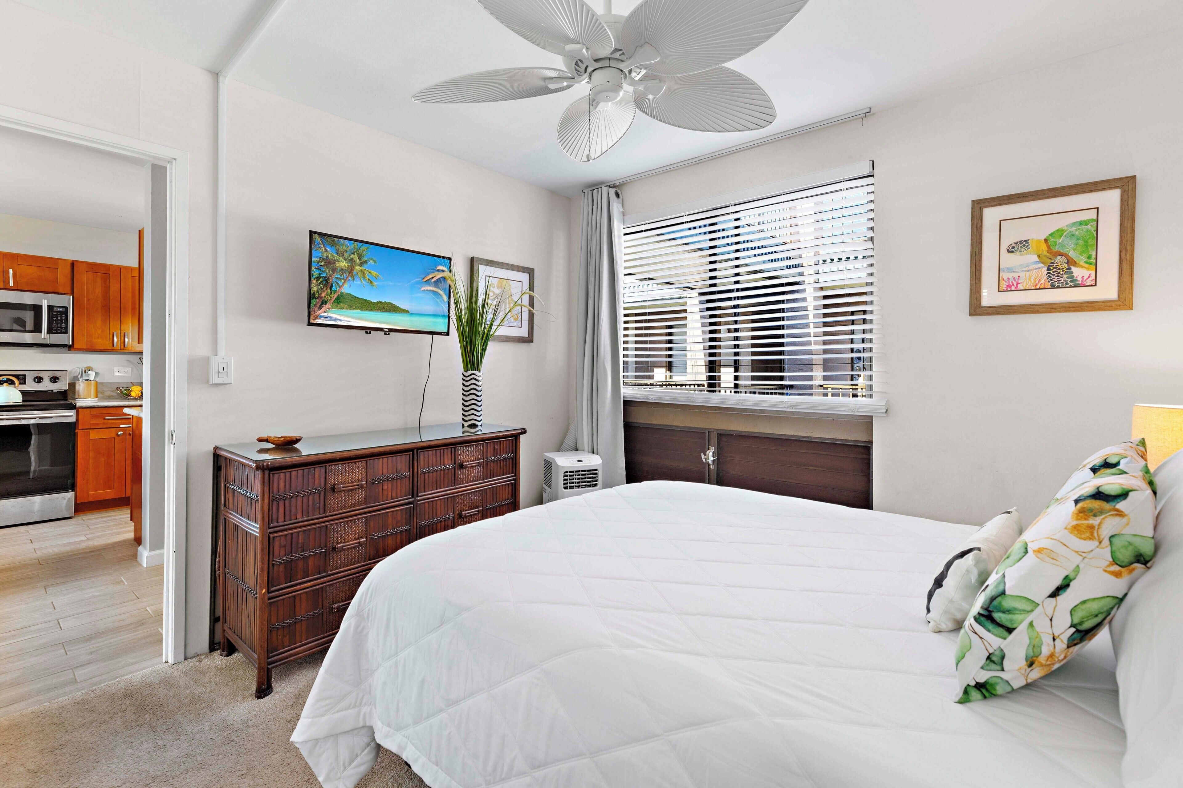 A bedroom with a bed, a ceiling fan, a window with blinds, a TV, and a kitchen visible through an open door.