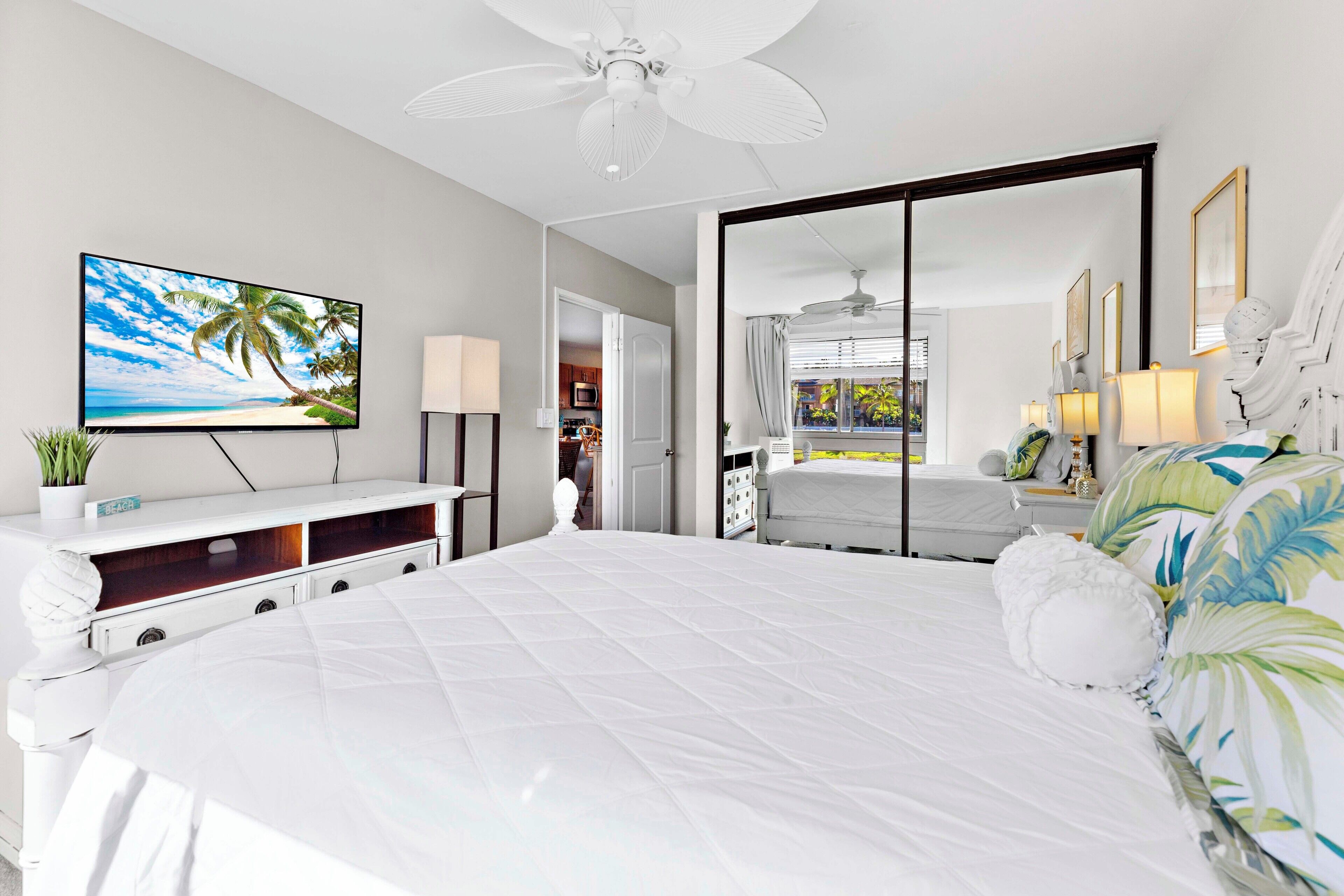 A modern bedroom with a large bed, a TV on a white console, a ceiling fan, and a large mirror.