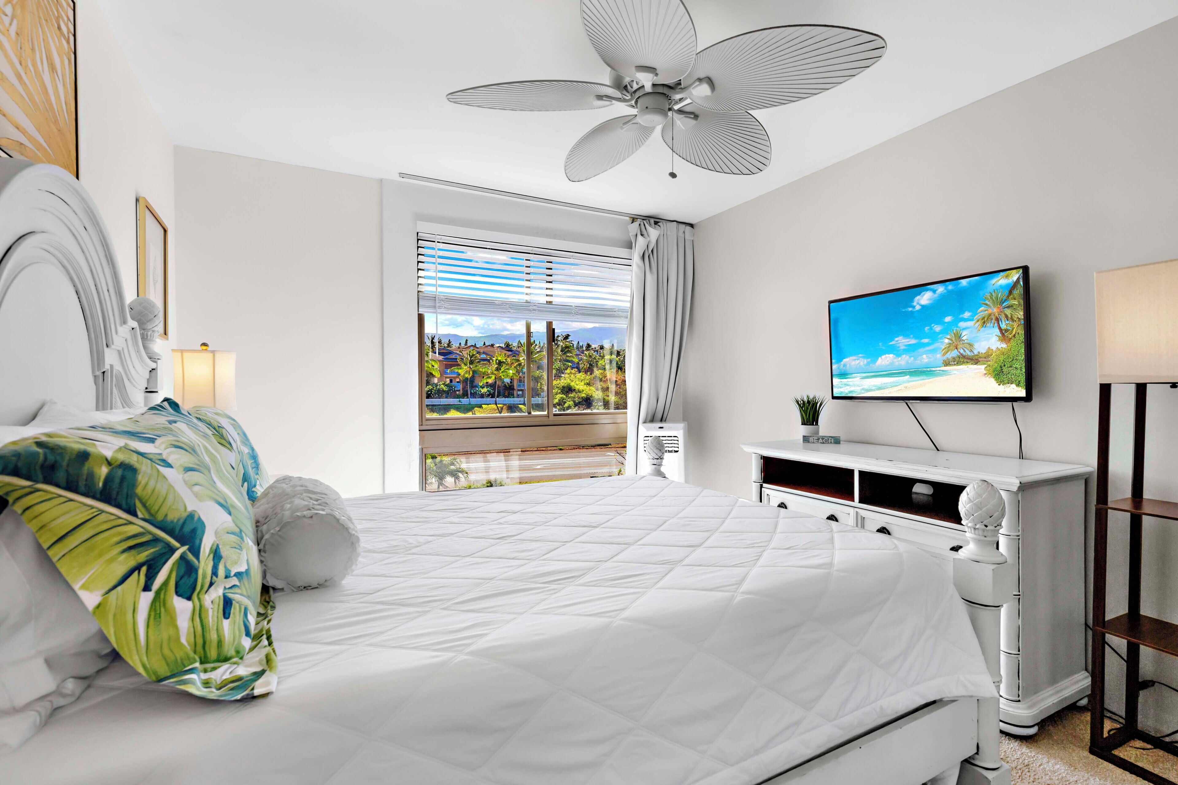 Un dormitorio moderno con una cama grande, ventilador de techo, televisión y vistas a la playa a través de la ventana.