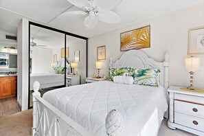 Une chambre à coucher avec un lit blanc, une table de chevet avec une lampe, un ventilateur de plafond et un grand miroir.