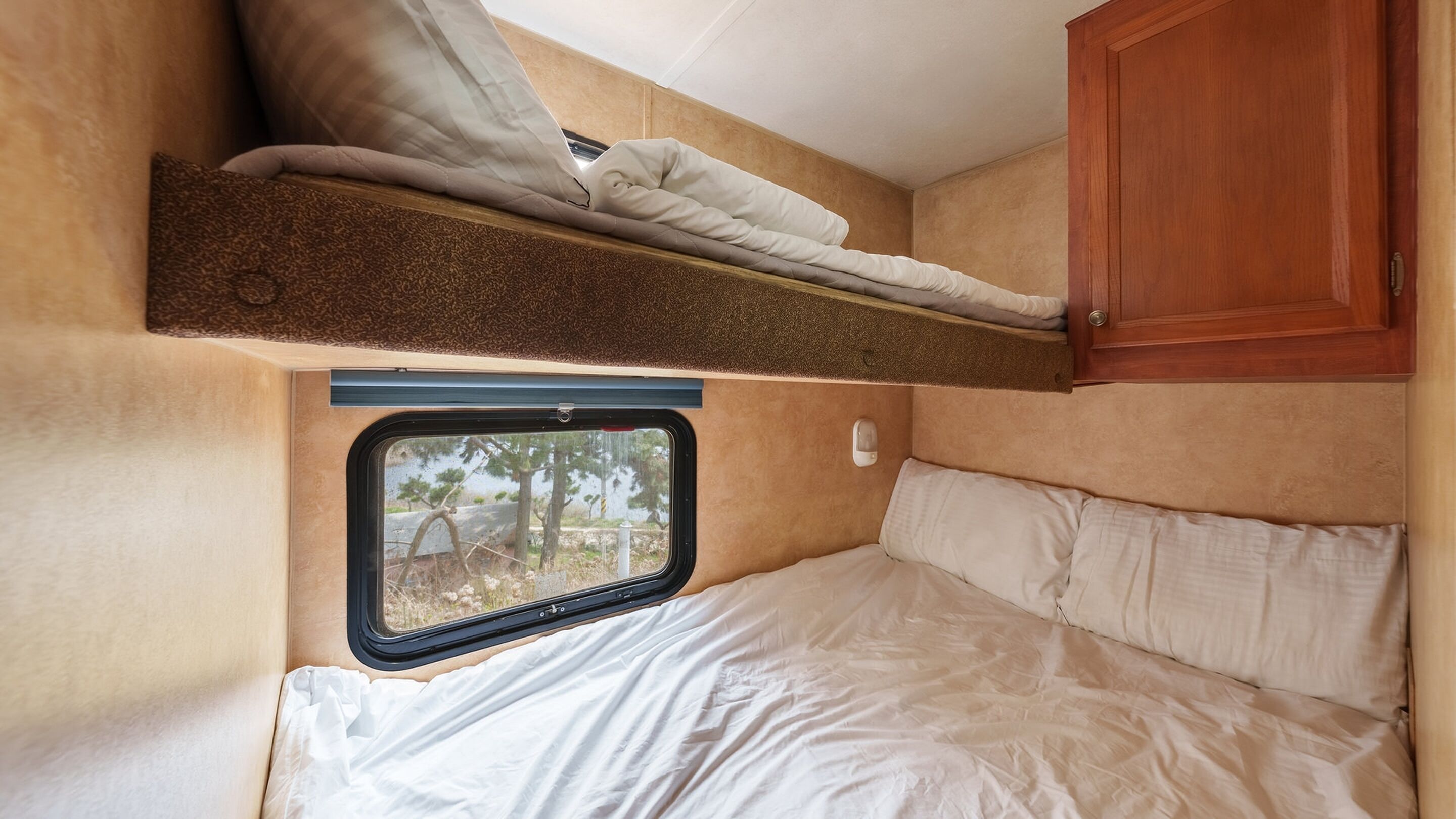 A compact RV interior with a bunk bed, a small window, and a wooden cabinet.
