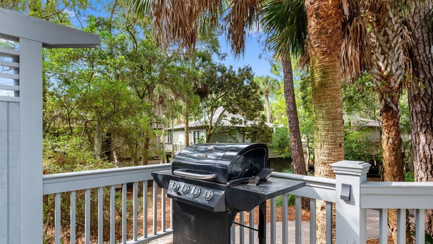 A barbecue grill on a wooden deck with a white railing, surrounded by trees and a house in the background. - Relaxing Coastal Stay with Shared Pool | La Mer (Johns Island)