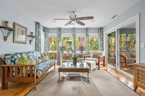 Ein geräumiges Wohnzimmer mit einem großen Glasfenster, das zu einem Außenbereich führt, einem Deckenventilator und verschiedenen Sitzgelegenheiten, darunter ein Sofa und Sessel.