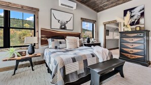 Un dormitorio con una cama grande, cabecera de madera, cómoda con cajones, mesita de noche, lámpara y ventana con vista a árboles.