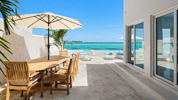 A wooden table and chairs with a beige umbrella, overlooking a beach with clear blue water. - Modern, Stunning & Bright Central Beach Villa (Nassau)