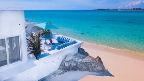A beachfront property with a white building, a blue outdoor seating area with a table and chairs, and a view of the ocean. - Modern, Stunning & Bright Central Beach Villa (Nassau)