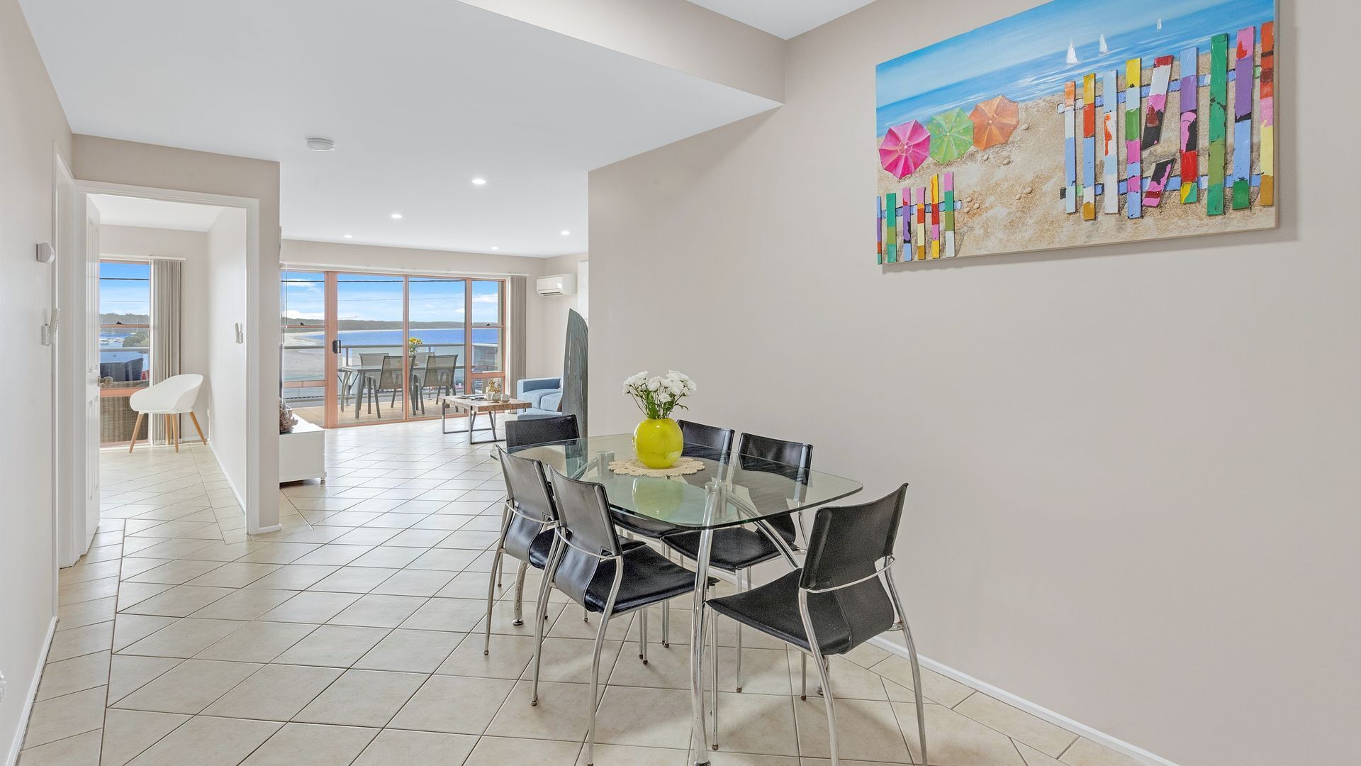 A dining area with a glass table and chairs, a yellow vase with flowers, and a beach-themed painting on the wall.