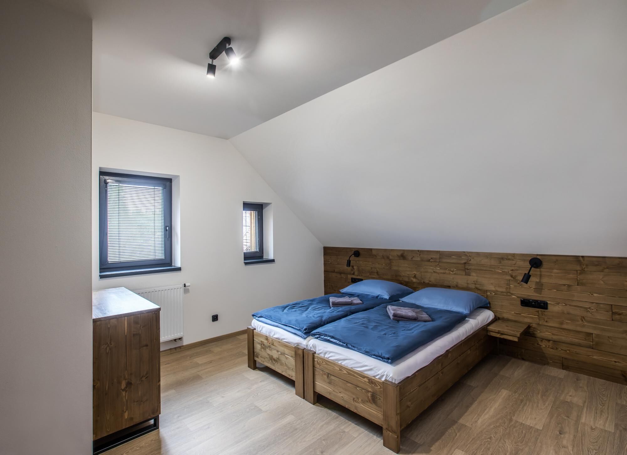A bedroom with a sloped ceiling, a wooden headboard, two beds with blue bedding, a small window, and a radiator.