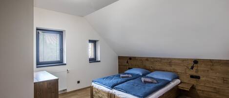 A bedroom with a sloped ceiling, a wooden headboard, two beds with blue bedding, a small window, and a radiator.
