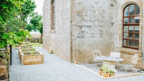 Un espace extérieur aménagé dans un style rustique, avec des jardinières en bois et des chaises en métal, un mur en pierre et une grande fenêtre en arc de cercle.