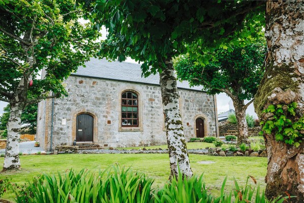 Un bâtiment en pierre, doté d’une porte sombre et de fenêtres en arc de cercle, entouré de verdure et d’arbres.