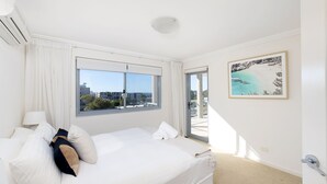 A hotel room with a bed, a window with a city view, a painting of a beach, and an air conditioning unit.