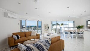 A modern living room with a brown sofa, white coffee table, and a dining area with white chairs and a wooden table.