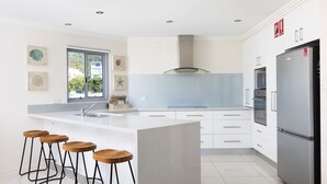 A modern kitchen with white cabinetry, a large island with stools, a stainless steel refrigerator, and a window with a view of buildings outside.
