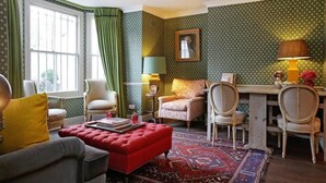 Ein gemütliches Wohnzimmer mit einem roten Hocker, zwei Sesseln, einem kleinen Tisch und einem gemusterten Teppich.