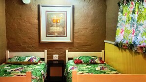 Une chambre dotée de deux lits simples, chacun recouvert d’une couvre-lit ornée de motifs tropicaux, avec un tableau encadré au mur et un rideau à motifs floraux.