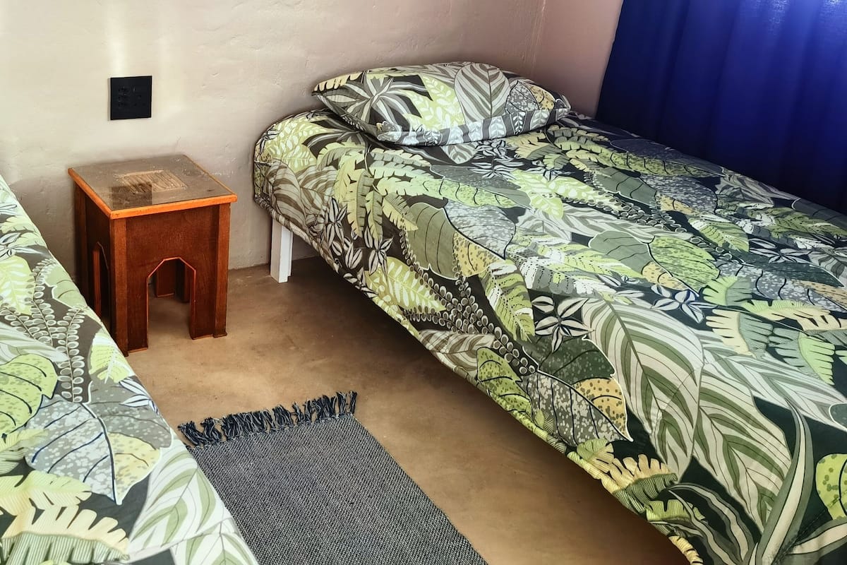 Una habitación con dos camas individuales, cada una con colcha estampada de hojas, una mesita de noche de madera y una cortina azul oscuro.