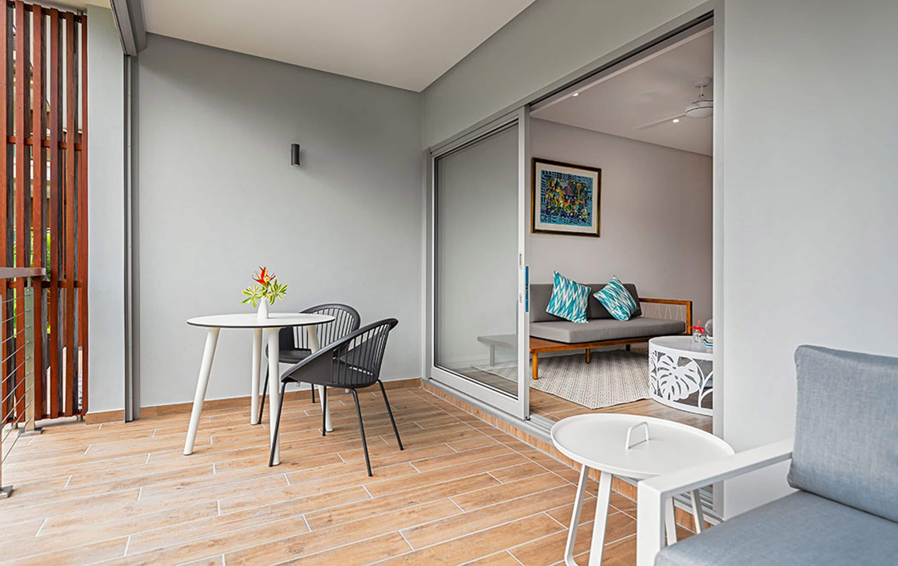 En modern uteplats med ett runt bord och stolar, en soffa och en skjutdörr i glas som leder in till ett inre utrymme med en soffa och kuddar.
