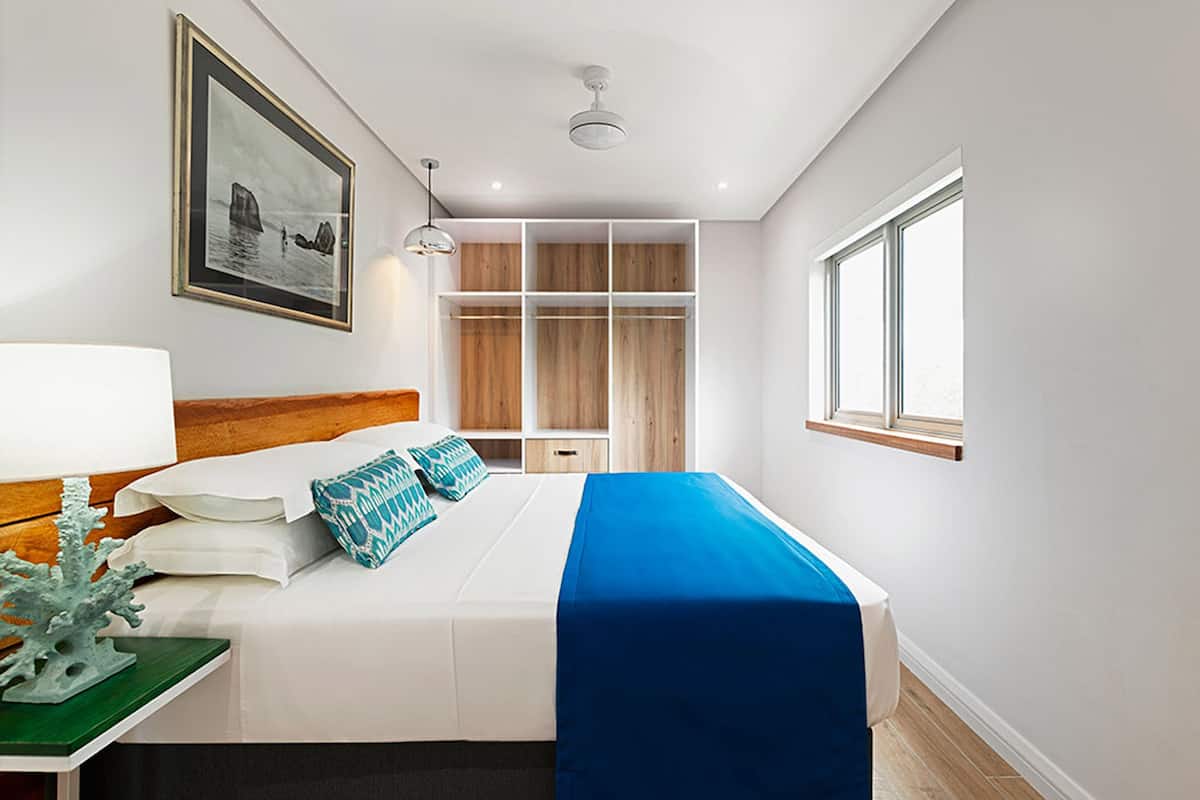 A modern hotel room with a large bed, a wooden headboard, a built-in wardrobe, a bedside table with a lamp, and a framed picture on the wall.