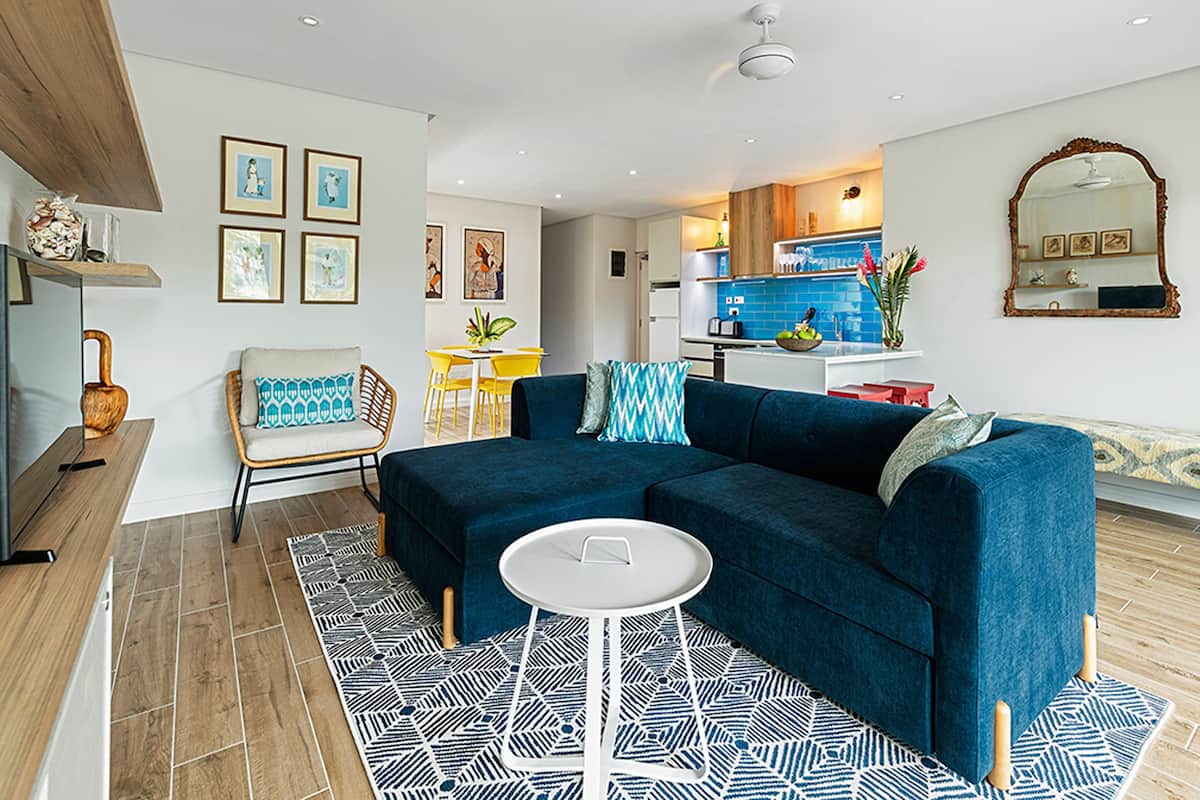 A modern living room with a blue sectional sofa, a white coffee table, and a patterned rug. There is a dining area with yellow chairs and a kitchen in the background.