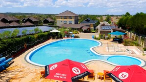 A swimming pool with red umbrellas and lounge chairs, surrounded by houses and solar panels.